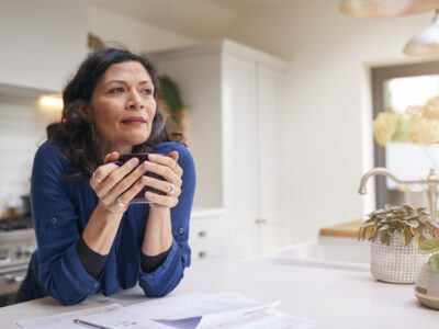 Thoughtful Mature Woman Reviewing Domestic Finances And Paperwork Drinking Coffee In Kitchen At Home