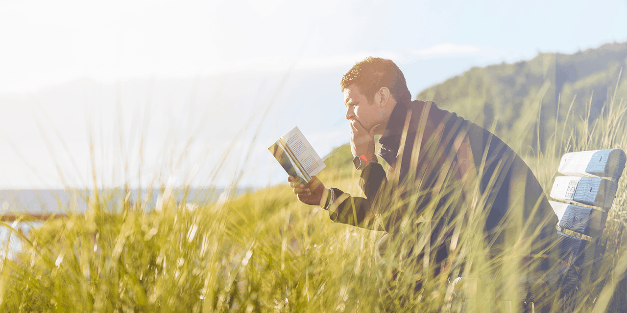 Man sitting on a bench in a field reading a book about investing