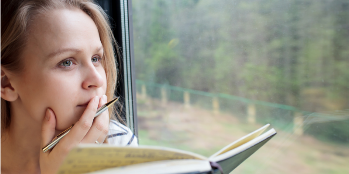 Young woman on a train writing notes in a diary or journal staring thoughtfully out of the window with her pen to her lips as she thinks of what to write